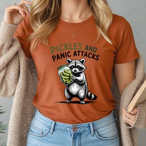 May include: Rust-coloured t-shirt with a raccoon holding a jar of pickles. The text "Pickles and Panic Attacks" is above the raccoon. The raccoon is black, white, and grey, with a striped tail. The pickles are green.