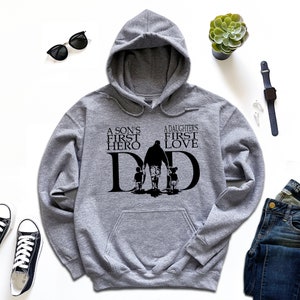 May include: A gray hooded sweatshirt with the text "A son's first hero, A daughter's first love, DAD" in black. The text is surrounded by a silhouette of a father walking with two children.