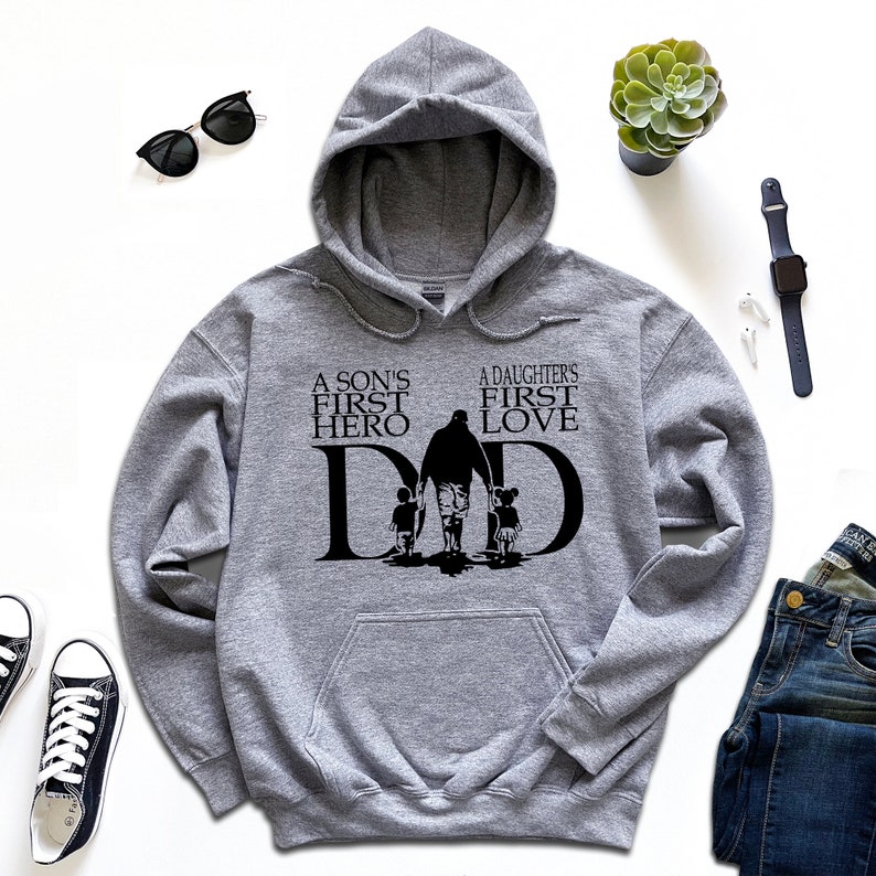 May include: A gray hooded sweatshirt with the text "A son's first hero, A daughter's first love, DAD" in black. The text is surrounded by a silhouette of a father walking with two children.