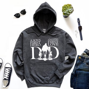May include: A dark gray hooded sweatshirt with a white graphic design. The graphic features a silhouette of a father walking with a son and daughter, with the text "A son's first hero" and "A daughter's first love" above the silhouette. The text "DAD" is incorporated into the silhouette.