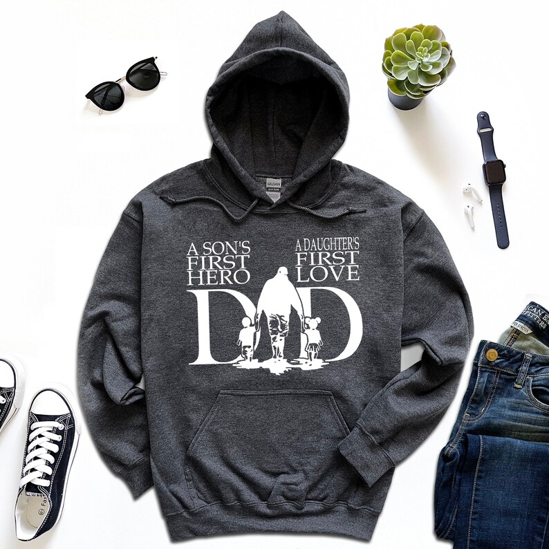 May include: A dark gray hooded sweatshirt with a white graphic design. The graphic features a silhouette of a father walking with a son and daughter, with the text "A son's first hero" and "A daughter's first love" above the silhouette. The text "DAD" is incorporated into the silhouette.