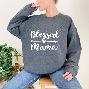 Puede incluir: Una sudadera gris con el texto "Blessed Mama" en letras blancas con un gr&aacute;fico de flecha y coraz&oacute;n.