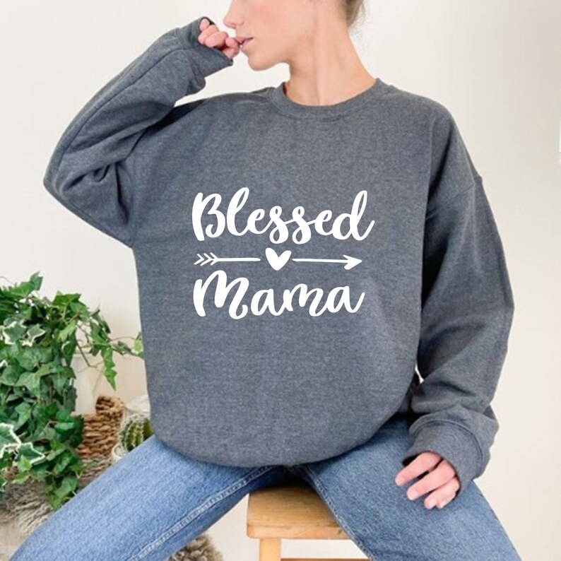 Puede incluir: Una sudadera gris con el texto "Blessed Mama" en letras blancas con un gr&aacute;fico de flecha y coraz&oacute;n.