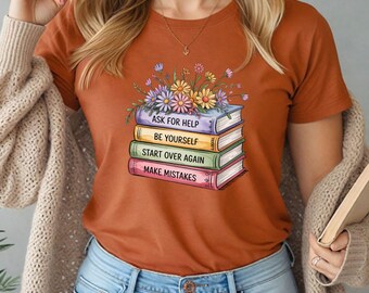 Puede incluir: Camiseta color óxido con una pila de libros y flores. Cada libro tiene un texto diferente: "ASK FOR HELP", "BE YOURSELF", "START OVER AGAIN" y "MAKE MISTAKES". La camiseta se combina con unos vaqueros azules.