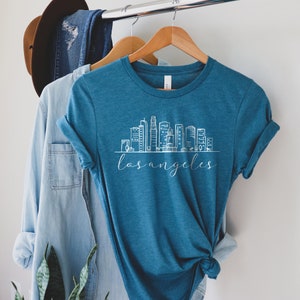 Peut inclure: Un t-shirt bleu sarcelle avec un graphique blanc de l'horizon de Los Angeles. Le t-shirt est suspendu &agrave; un cintre en bois.