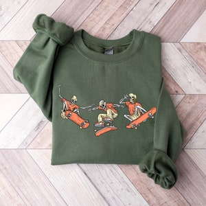 May include: Green sweatshirt with a graphic of three skeletons skateboarding. The skeletons are wearing orange shirts and hats. The graphic is in a cartoon style.