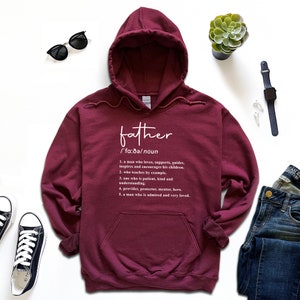 K&ouml;nnte beinhalten: Ein weinroter Kapuzenpullover mit dem Wort "father" und seiner Definition in wei&szlig;er Schrift. Die Definition beschreibt die Eigenschaften eines Vaters. Der Hoodie hat eine Kapuze und eine Vordertasche.
