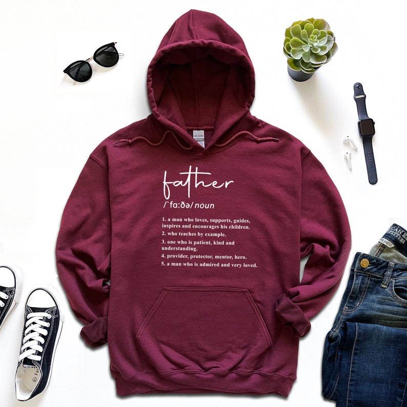K&ouml;nnte beinhalten: Ein weinroter Kapuzenpullover mit dem Wort "father" und seiner Definition in wei&szlig;er Schrift. Die Definition beschreibt die Eigenschaften eines Vaters. Der Hoodie hat eine Kapuze und eine Vordertasche.