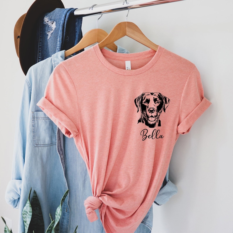May include: A coral pink t-shirt with a black silhouette of a Labrador Retriever and the name "Bella" printed on the front.