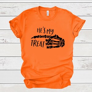May include: Orange t-shirt with a black skeleton hand pointing to the right and the text "He's my treat".