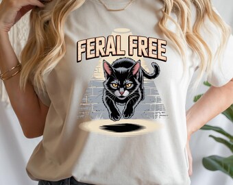 Puede incluir: Camiseta color crema con un gráfico de un gato negro con ojos amarillos, caminando hacia el espectador. El texto "FERAL FREE" está encima del gato en una fuente estilizada. El gato está sobre un fondo de pared de ladrillos.