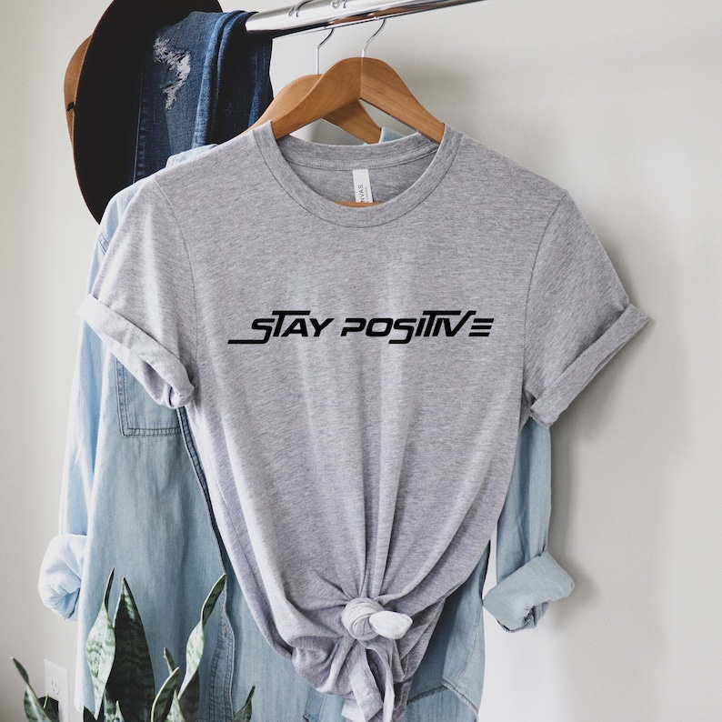 Puede incluir: Camiseta gris con texto negro que dice "STAY POSITIVE" en una fuente estilizada.