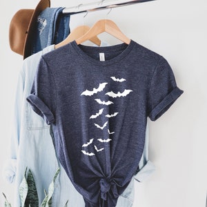 Puede incluir: Una camiseta azul oscuro con un gr&aacute;fico blanco de murci&eacute;lagos volando en l&iacute;nea vertical. La camiseta est&aacute; colgada en una percha de madera.