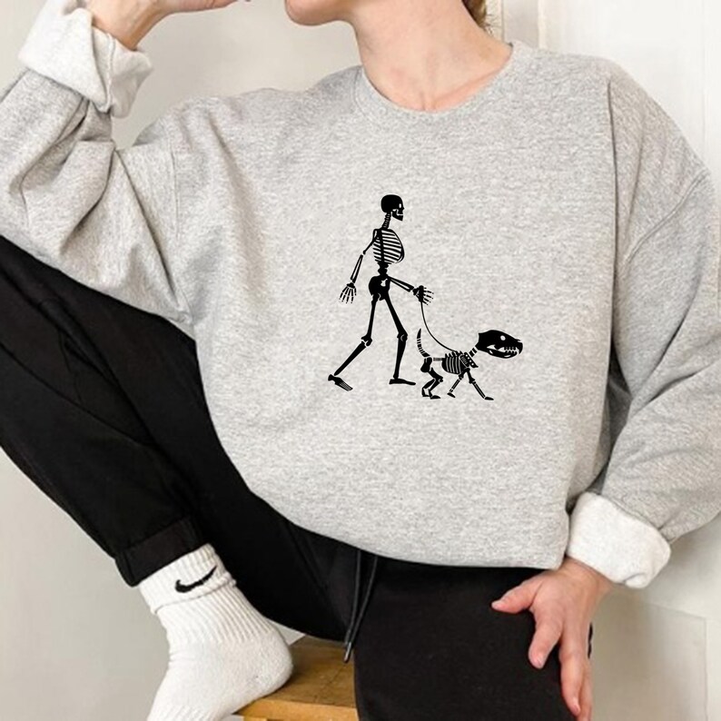 Pode incluir: Uma camisa de moletom cinza com um gr&aacute;fico de esqueleto preto de um esqueleto passeando um cachorro esqueleto.