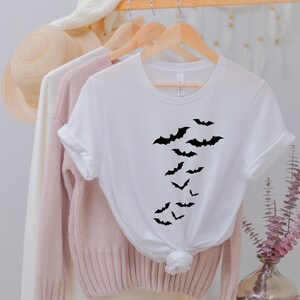 Puede incluir: Camiseta blanca con una silueta negra de murci&eacute;lagos volando en l&iacute;nea vertical en la parte delantera.