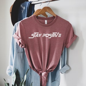 Puede incluir: Una camiseta de color malva con el texto "STAY POSITIVE" en letras blancas. La camiseta est&aacute; colgada de una percha de madera.