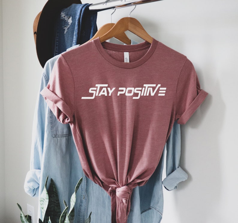 Puede incluir: Una camiseta de color malva con el texto "STAY POSITIVE" en letras blancas. La camiseta est&aacute; colgada de una percha de madera.