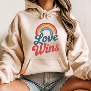 Op de afbeelding: Een beige hoodie met een regenboogafbeelding en de woorden "Love Wins" in blauw en rood. De hoodie heeft een capuchon met trekkoord en een voorzak. Het ontwerp is gecentreerd op de voorkant.