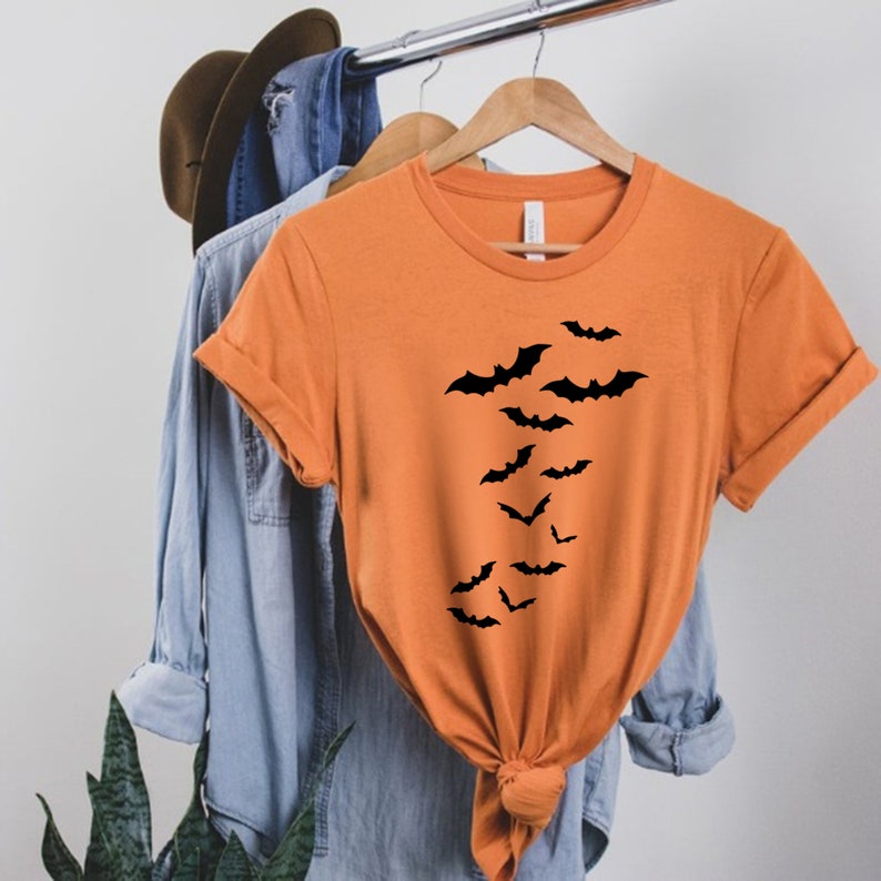 Puede incluir: Camiseta naranja con un dise&ntilde;o de murci&eacute;lago negro. Los murci&eacute;lagos vuelan en diagonal de arriba a la izquierda a abajo a la derecha de la camiseta.