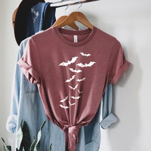 Puede incluir: Una camiseta de color marr&oacute;n rojizo con un gr&aacute;fico blanco de murci&eacute;lagos volando en bandada. La camiseta est&aacute; anudada a la cintura y est&aacute; colgada en una percha.
