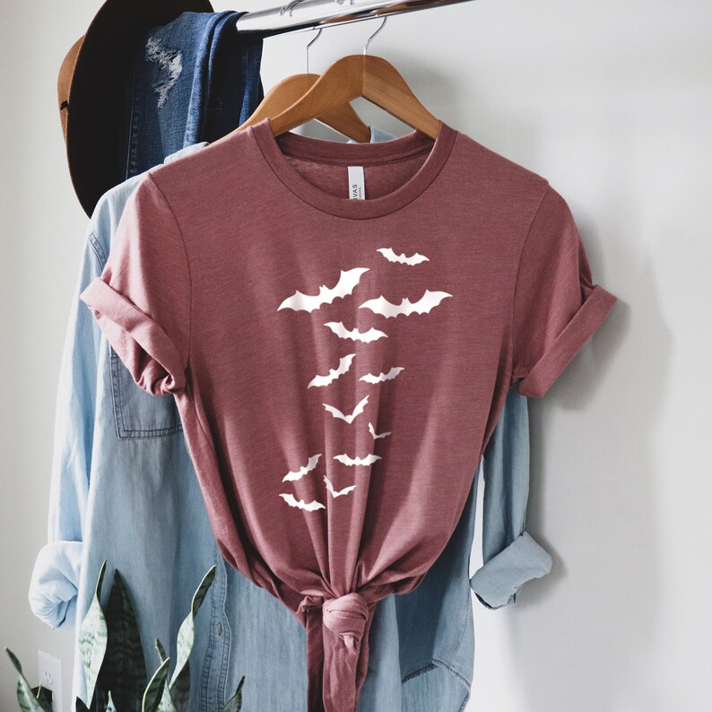 Puede incluir: Una camiseta de color marr&oacute;n rojizo con un gr&aacute;fico blanco de murci&eacute;lagos volando en bandada. La camiseta est&aacute; anudada a la cintura y est&aacute; colgada en una percha.
