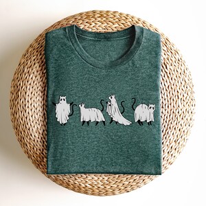 May include: A dark green t-shirt with a graphic of five cats dressed as ghosts. The cats are in a line, facing forward, and each has a white ghost sheet draped over their body. The graphic is in black and white.