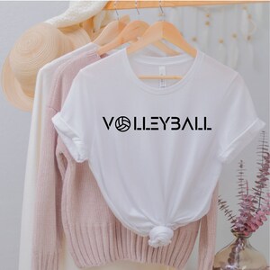 May include: White t-shirt with black text that reads "VOLLEYBALL" with a volleyball graphic in the middle of the word.