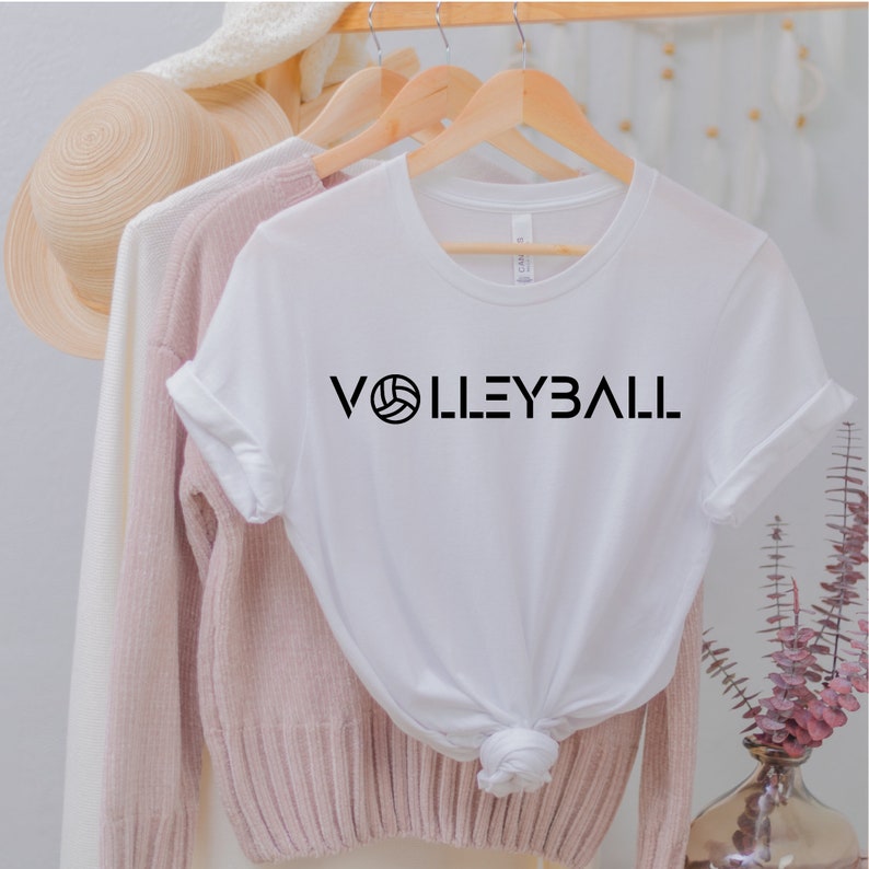 May include: White t-shirt with black text that reads "VOLLEYBALL" with a volleyball graphic in the middle of the word.