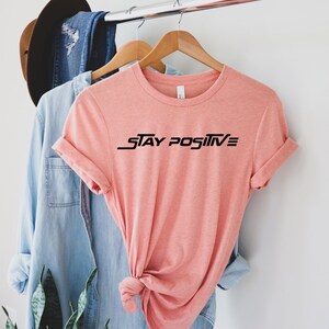 Puede incluir: Una camiseta rosa con el texto "STAY POSITIVE" en letras negras.