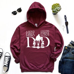 May include: Burgundy hooded sweatshirt with a white graphic design. The graphic features a father walking with a son and daughter, with the text "A son's first hero" and "A daughter's first love" above the image. The word "DAD" is incorporated into the design.