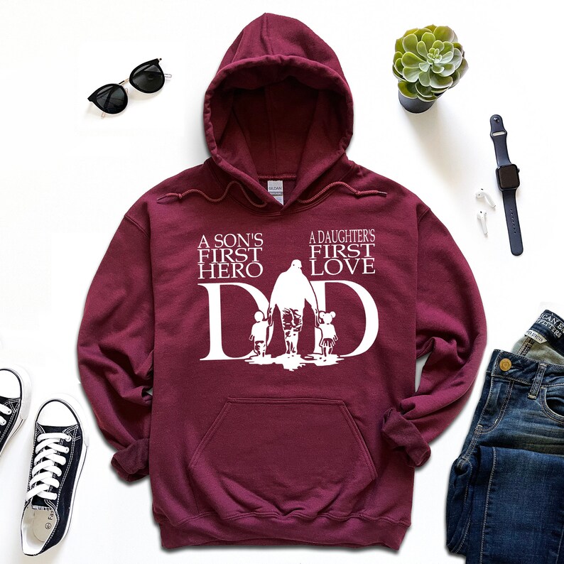 May include: Burgundy hooded sweatshirt with a white graphic design. The graphic features a father walking with a son and daughter, with the text "A son's first hero" and "A daughter's first love" above the image. The word "DAD" is incorporated into the design.