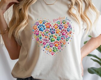Puede incluir: Camiseta color crema con un diseño en forma de corazón compuesto por huellas de patas coloridas. Las palabras "Pawfect Love" están escritas encima del corazón. La camiseta está hecha de un material suave.