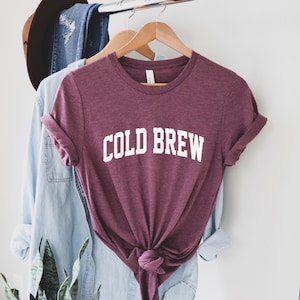May include: A maroon t-shirt with the text "COLD BREW" in white letters. The shirt is knotted at the bottom and is hanging on a hanger.