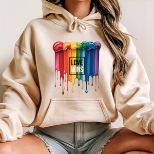 May include: A beige hoodie featuring a colorful rainbow design with dripping paint effect. The design includes the text "LOVE WINS" in a black square. The hoodie has a front pocket and a drawstring hood.