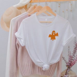 以下が含まれることがあります： 左胸に小さなオレンジ色の幽霊のグラフィックが描かれた白いTシャツ。シャツは、ピンクのセーターと麦わら帽子と一緒にハンガーにかけられています。