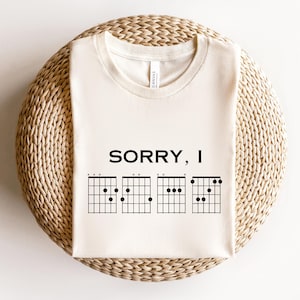 Puede incluir: Camiseta color crema con el texto "SORRY, I" sobre cuatro diagramas de acordes de guitarra negros. La camiseta est&aacute; hecha de un material suave y se muestra sobre una superficie circular tejida. Es una prenda de vestir con tem&aacute;tica musical.