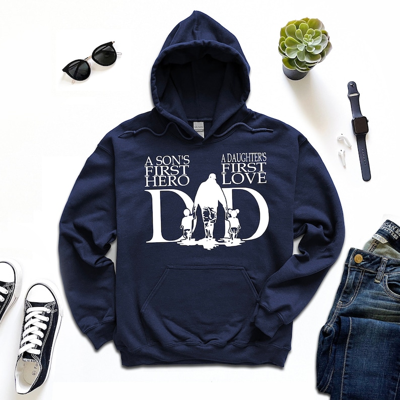 May include: Navy blue hooded sweatshirt with a white graphic design. The graphic features a father walking with a son and daughter, with the text "A son's first hero" and "A daughter's first love" above the figures. The word "DAD" is incorporated into the design.