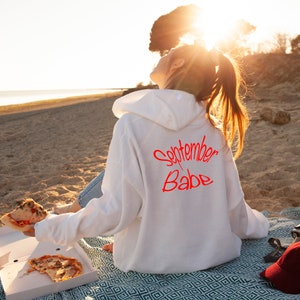 Puede incluir: Una sudadera con capucha blanca con el texto "September Babe" escrito en rojo en la espalda. La persona que lleva la sudadera est&aacute; sentada en una manta en una playa.