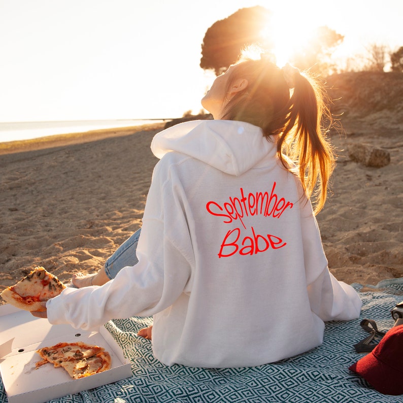 Puede incluir: Una sudadera con capucha blanca con el texto "September Babe" escrito en rojo en la espalda. La persona que lleva la sudadera est&aacute; sentada en una manta en una playa.