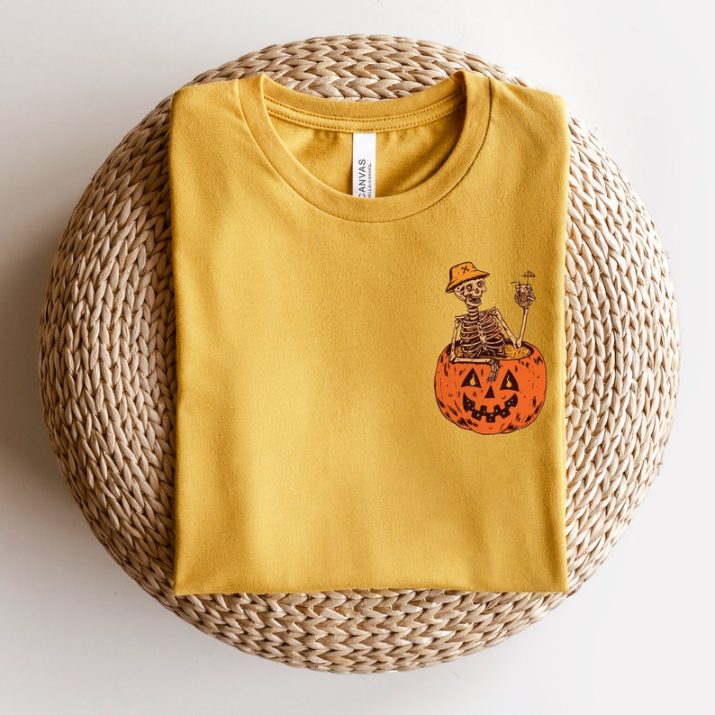 May include: A mustard yellow t-shirt with a graphic of a skeleton sitting in a jack-o'-lantern holding a drink.