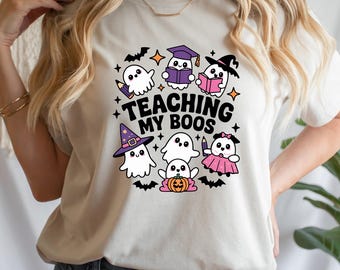 May include: A cream-coloured t-shirt with a Halloween graphic. The design features cartoon ghosts, bats, stars, and the text "TEACHING MY BOOS". The ghosts are wearing hats and holding books and pencils.