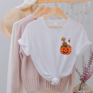 May include: White t-shirt with a graphic of a skeleton sitting inside a jack-o'-lantern. The skeleton is wearing a hat and holding a cigarette.