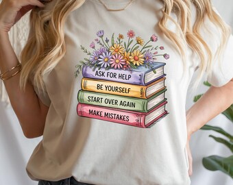 Puede incluir: Camiseta color crema con un diseño de libros apilados y flores. Los libros son de varios colores y tienen las frases "ASK FOR HELP", "BE YOURSELF", "START OVER AGAIN" y "MAKE MISTAKES". El arreglo floral incluye flores moradas, amarillas y rosas.