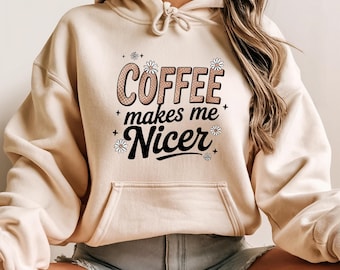 May include: A beige hoodie with a front pocket and drawstring. The front of the hoodie features the text "COFFEE makes me Nicer" in a mix of fonts and colors, with floral accents.