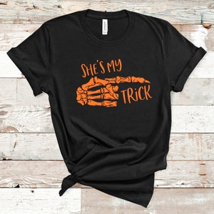 May include: Black t-shirt with an orange skeleton hand pointing to the right and the text "She's my trick" printed on it.