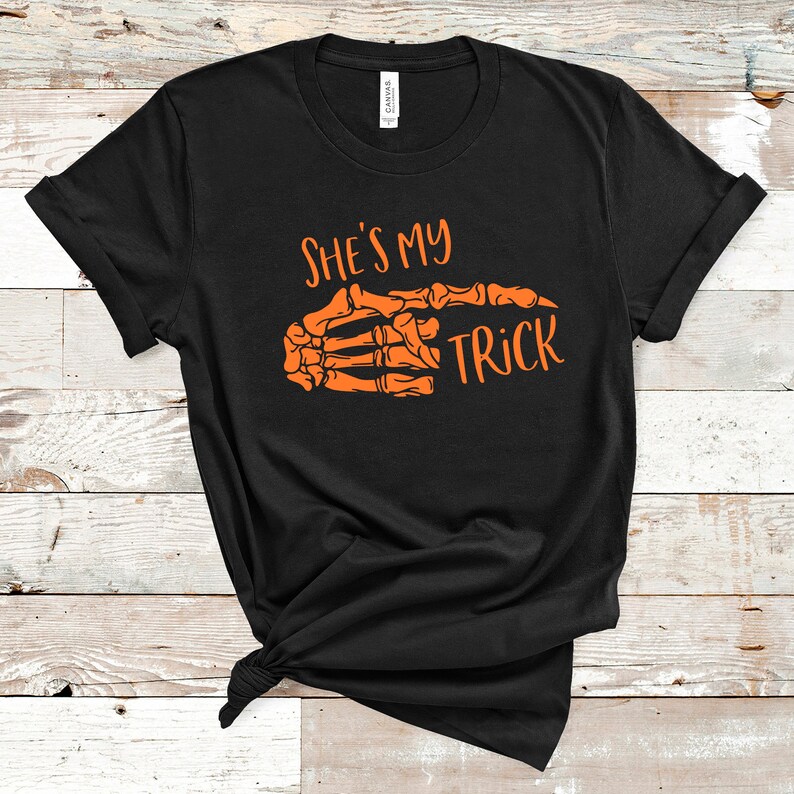 May include: Black t-shirt with an orange skeleton hand pointing to the right and the text "She's my trick" printed on it.