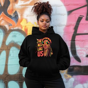 May include: Black hoodie featuring a graphic of a woman wearing star-shaped sunglasses and a colorful head wrap. The text "THEY NOT LIKE US" is printed on the front. The hoodie has a drawstring hood and a kangaroo pocket.