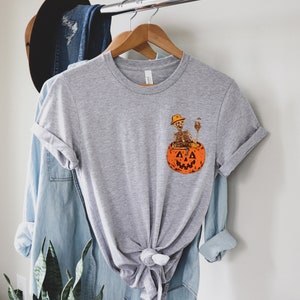 May include: A gray t-shirt with a graphic of a skeleton sitting in a jack-o'-lantern. The skeleton is wearing a hat and holding a drink.