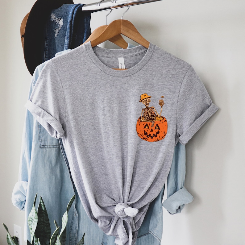 May include: A gray t-shirt with a graphic of a skeleton sitting in a jack-o'-lantern. The skeleton is wearing a hat and holding a drink.