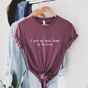 Puede incluir: Una camiseta de color granate con el texto "i put my book down to be here." impreso en blanco.